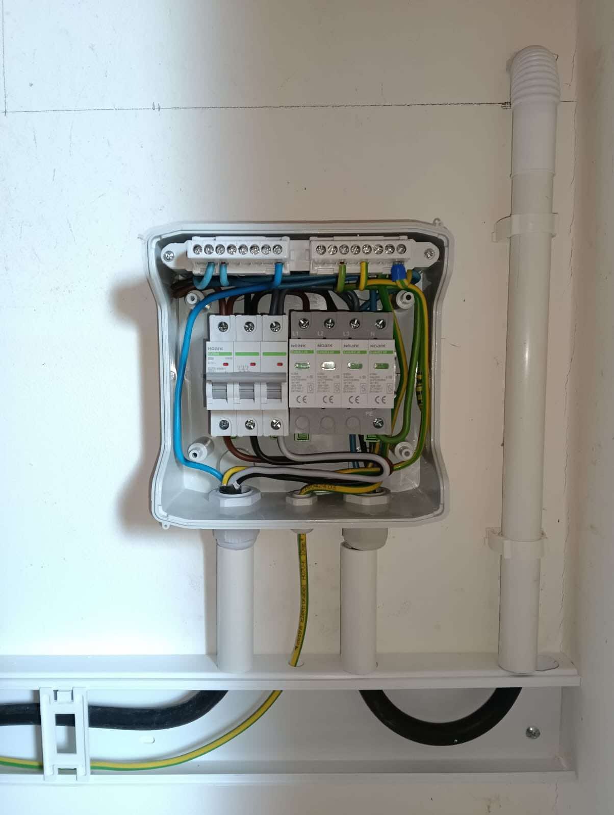 Otwarta skrzynka elektryczna z zainstalowanymi bezpiecznikami i przewodami w kolorach niebieskim, brązowym, żółto-zielonym, umieszczona na białej ścianie z widocznymi rurkami PCV.