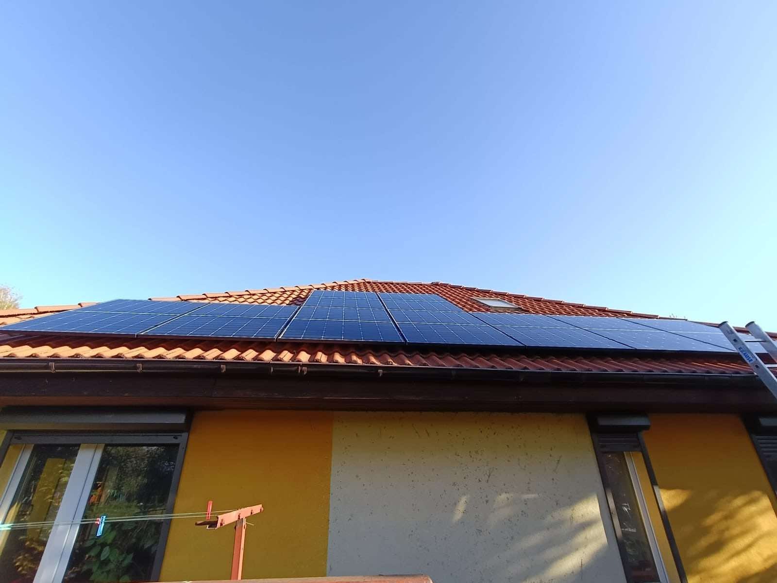 Panel słoneczny na dachu z czerwonej dachówki, z widoczną drabiną opartą o rynnę i fragmentem elewacji budynku w odcieniach żółci i beżu.