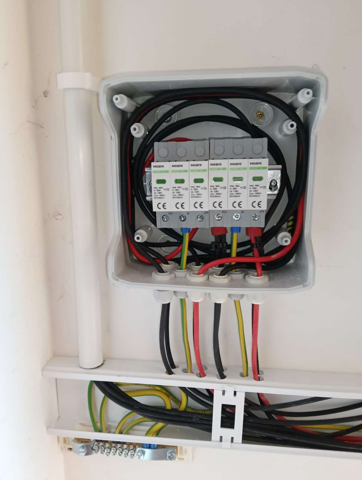 Rozdzielnica elektryczna z zabezpieczeniami przepięciowymi Noark, widoczne przewody w kolorach czarnym, czerwonym, żółto-zielonym, zamontowana na białej ścianie.