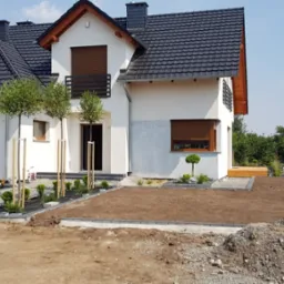 Nowoczesny dom jednorodzinny z czarną dachówką, drewnianą stolarką okienną i świeżo założonym ogrodem z młodymi drzewkami i rabatami z kostki brukowej.