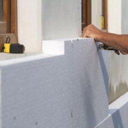 Blue Synergy - Pracownik docina nożem styropianowe płyty podczas ocieplania ściany budynku, obok okna leży miarka zwijana.