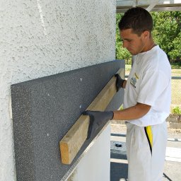 Blue Synergy - Pracownik w szarych rękawicach dociska deską szarą płytę izolacyjną do ściany z fakturą baranka podczas ocieplania elewacji budynku.