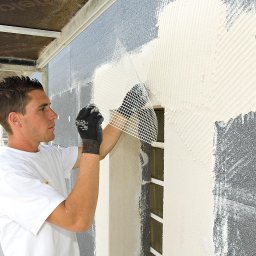 Blue Synergy - Pracownik w białej koszulce nakłada siatkę elewacyjną na ocieplony fragment ściany budynku, widoczny fragment rusztowania z lewej strony.