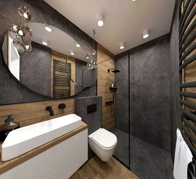 Nowoczesna łazienka z szarą, teksturowaną ścianą, białym blatem z umywalką nablatową, okrągłym lustrem z dekoracyjnymi lampami i kabiną prysznicową typu walk-in z czarną armaturą.