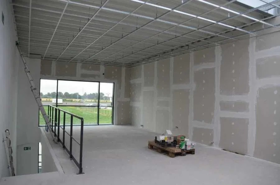 Wnętrze w trakcie wykończenia: ściany pokryte płytami gipsowo-kartonowymi, metalowa konstrukcja sufitu podwieszanego, drabina oparta o ścianę przy schodach, widok na zewnątrz przez duże okno.