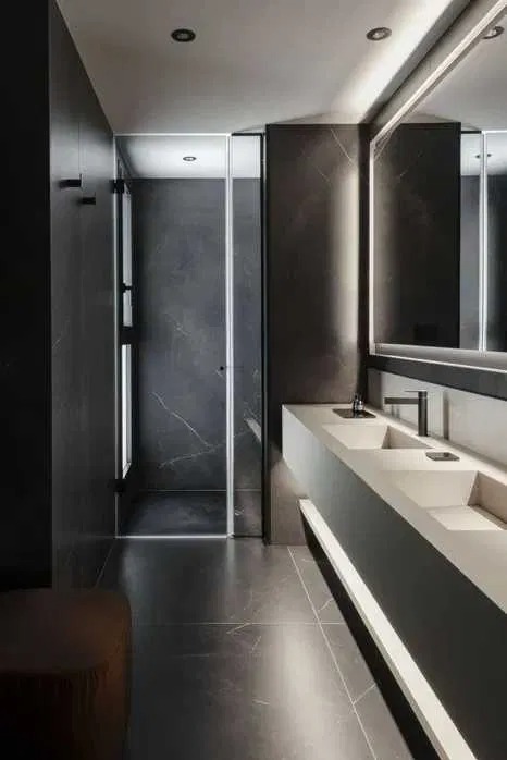 Nowoczesna łazienka z ciemnymi płytkami imitującymi marmur, podświetlaną umywalką z dwoma kranami i kabiną prysznicową ze szklanymi drzwiami.