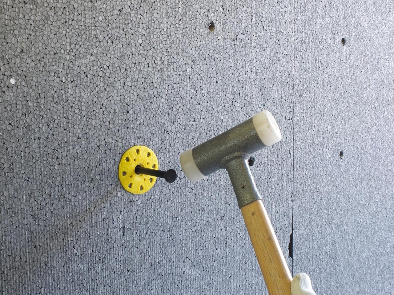 Przybijanie kołka mocującego izolację styropianową do ściany elewacyjnej za pomocą gumowego młotka. Widoczne otwory po wcześniejszych mocowaniach.