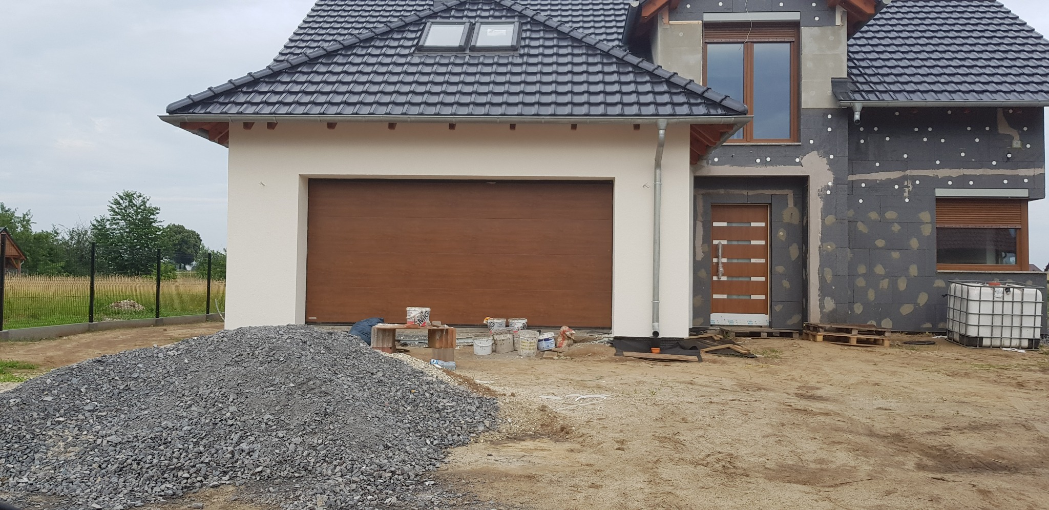 Dom w trakcie ocieplania, widoczne szare płyty styropianowe na elewacji, brązowa brama garażowa, okna z roletami, sterta gruzu i puszki po farbie.