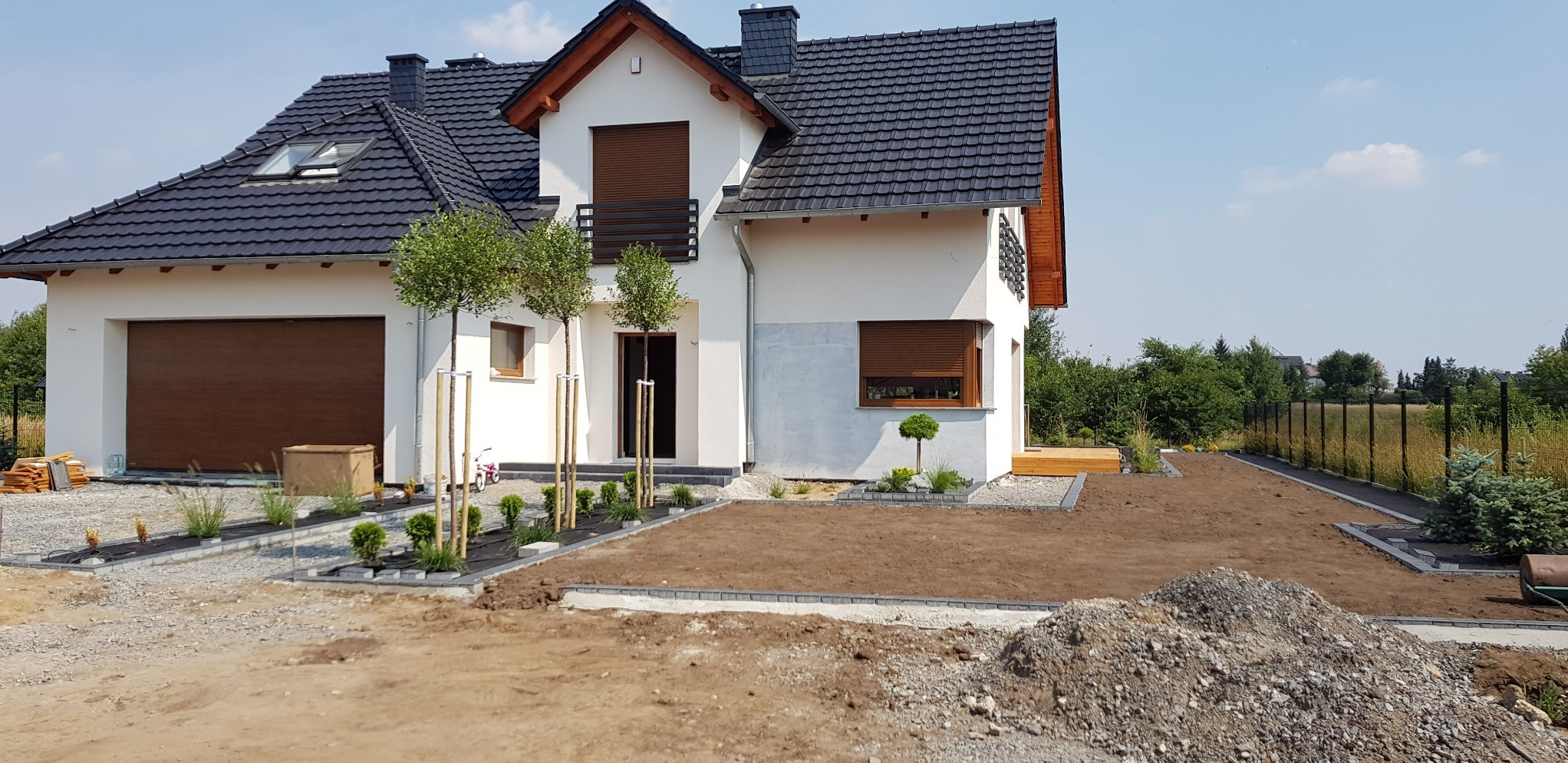 Nowoczesny dom jednorodzinny z czarną dachówką, drewnianą stolarką okienną i świeżo założonym ogrodem z młodymi drzewkami i rabatami z kostki brukowej.