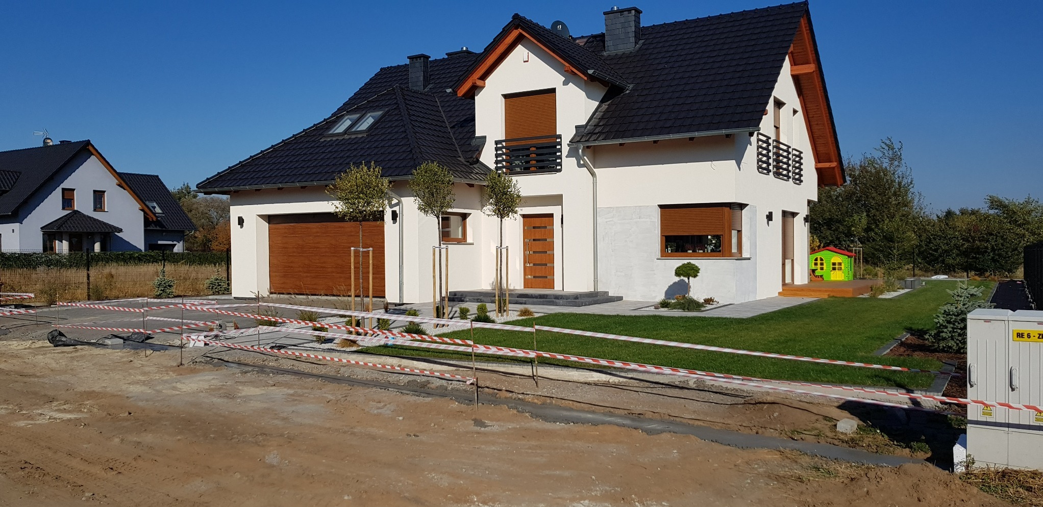 Nowoczesny dom jednorodzinny z czarnym dachem i jasną elewacją, z częściowo wykończonym podjazdem otoczonym taśmami ostrzegawczymi.