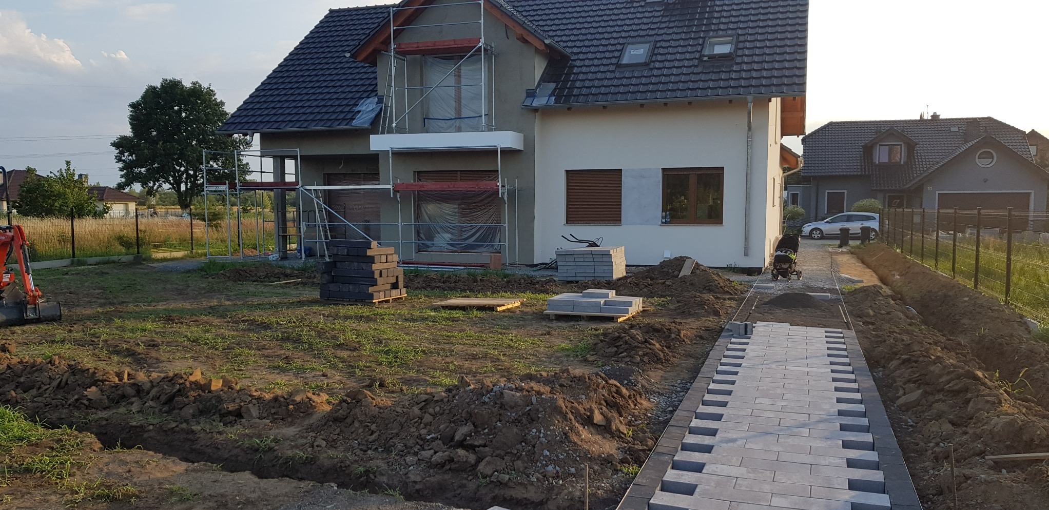 Dom jednorodzinny w trakcie prac wykończeniowych elewacji, widoczne rusztowania, ocieplone ściany oraz ułożony fragment chodnika z kostki brukowej, w tle drugi budynek i drzewa.