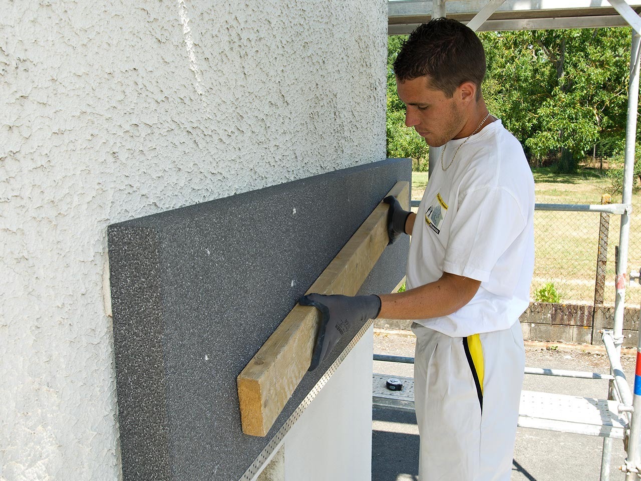 Pracownik w szarych rękawicach dociska deską szarą płytę izolacyjną do ściany z fakturą baranka podczas ocieplania elewacji budynku.