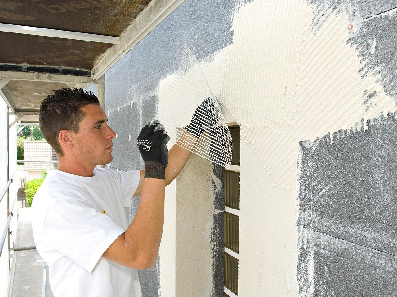 Pracownik w białej koszulce nakłada siatkę elewacyjną na ocieplony fragment ściany budynku, widoczny fragment rusztowania z lewej strony.