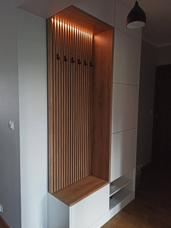 Biała szafa wnękowa z drewnianymi lamelami i podświetleniem LED, widoczne czarne wieszaki i półki, minimalistyczny design.