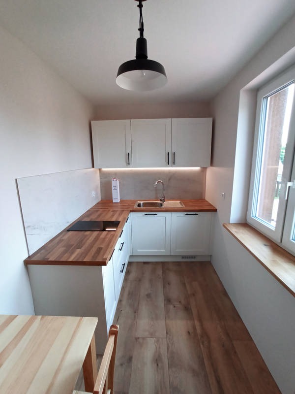 Jasna kuchnia w małym pomieszczeniu z drewnianym blatem, białymi szafkami i podłogą imitującą drewno, oświetlona lampą wiszącą i światłem dziennym z okna z drewnianym parapetem.