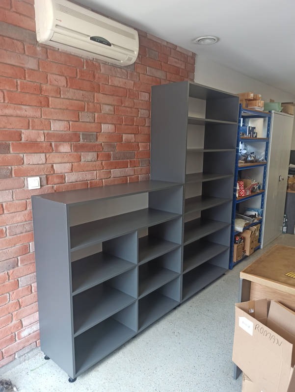 Szare regały z półkami na kółkach ustawione pod ścianą z czerwonej cegły, w tle widoczny klimatyzator i metalowy regał.