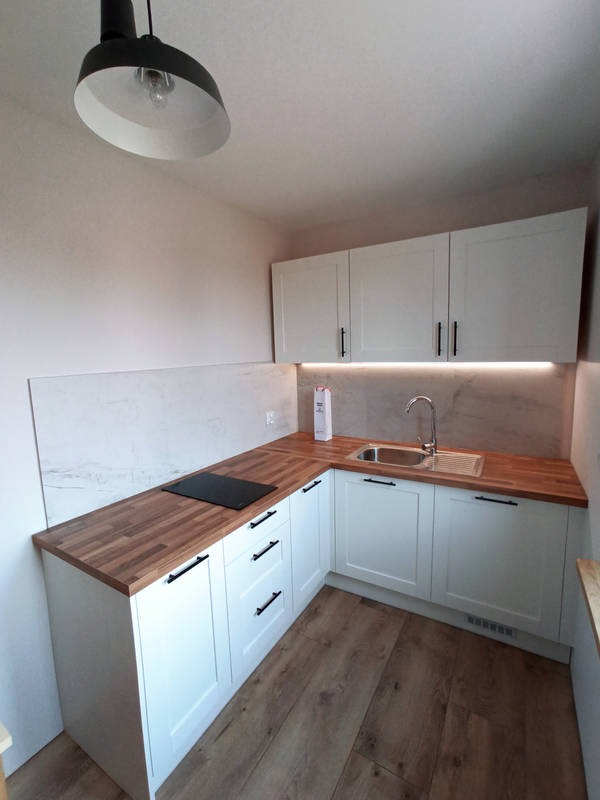 Nowoczesna, narożna zabudowa kuchenna z białymi frontami, drewnianym blatem i czarnymi uchwytami, oświetlona listwą LED pod szafkami wiszącymi, z płytą indukcyjną i zlewozmywakiem.