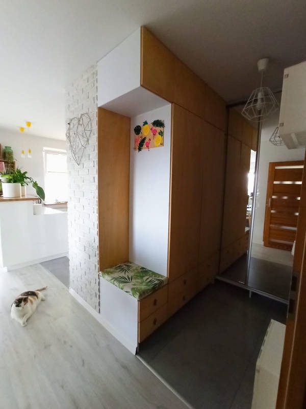 Wnęka z siedziskiem i szafa w przedpokoju, drewniane fronty, poduszka w tropikalne liście, na ścianie obrazek z owocami, kot leżący na podłodze.