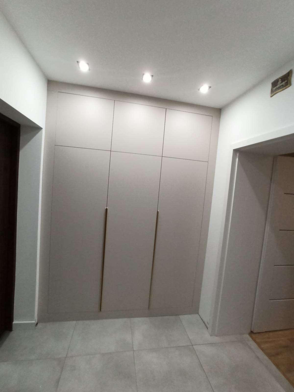 Trzydrzwiowa szafa wnękowa w odcieniach szarości z długimi, smukłymi uchwytami, wkomponowana w korytarz z szarą podłogą i oświetleniem punktowym.