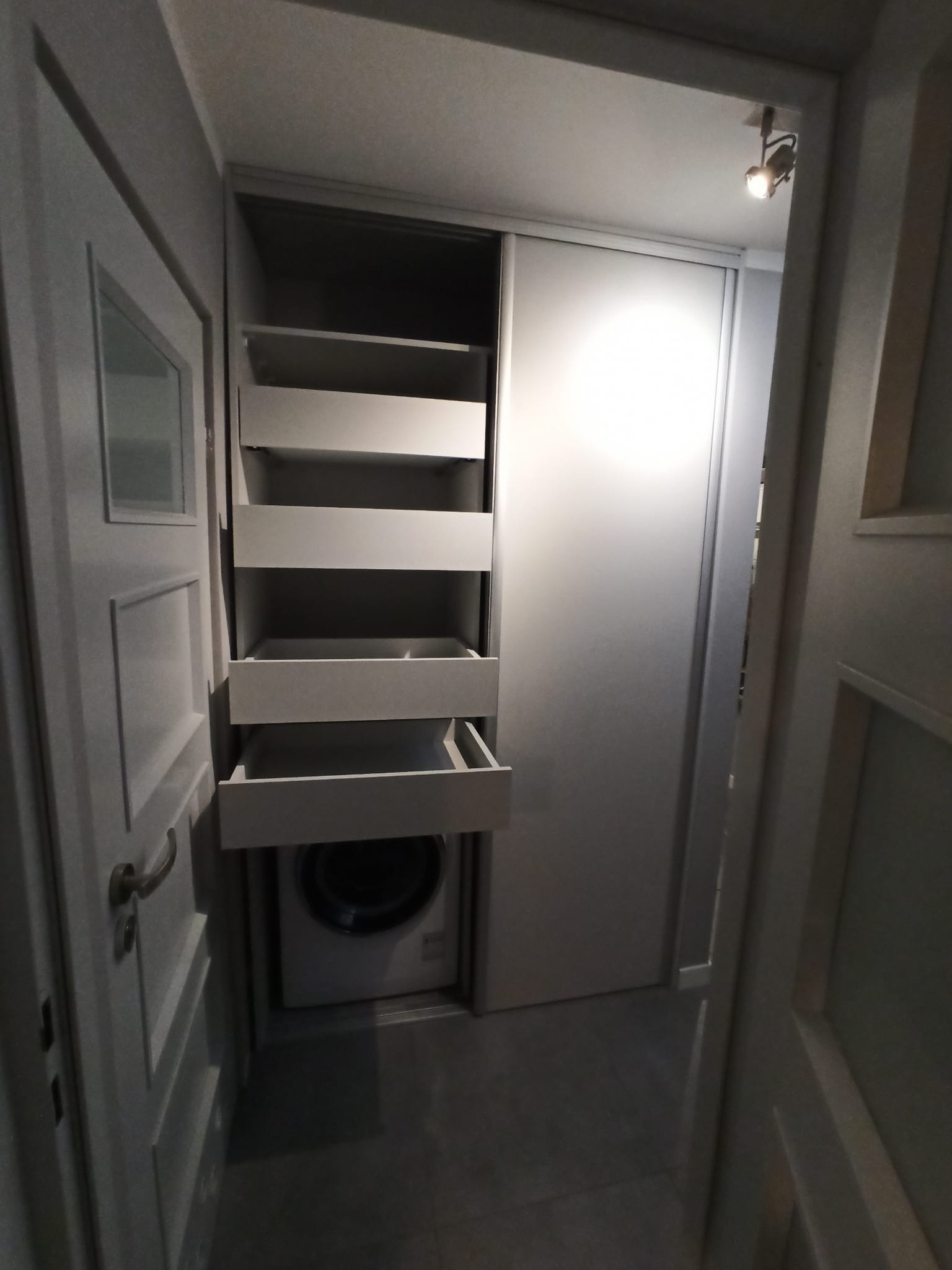 Szara szafa wnękowa z przesuwnymi drzwiami i otwartymi półkami, pod którymi umieszczona jest pralka, widoczna z perspektywy korytarza.