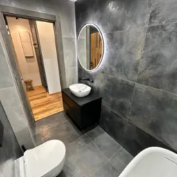 Nowoczesna łazienka z szarymi płytkami imitującymi beton, okrągłym lustrem z podświetleniem LED, białą umywalką nablatową na czarnej szafce i częściowo widoczną wanną oraz toaletą.