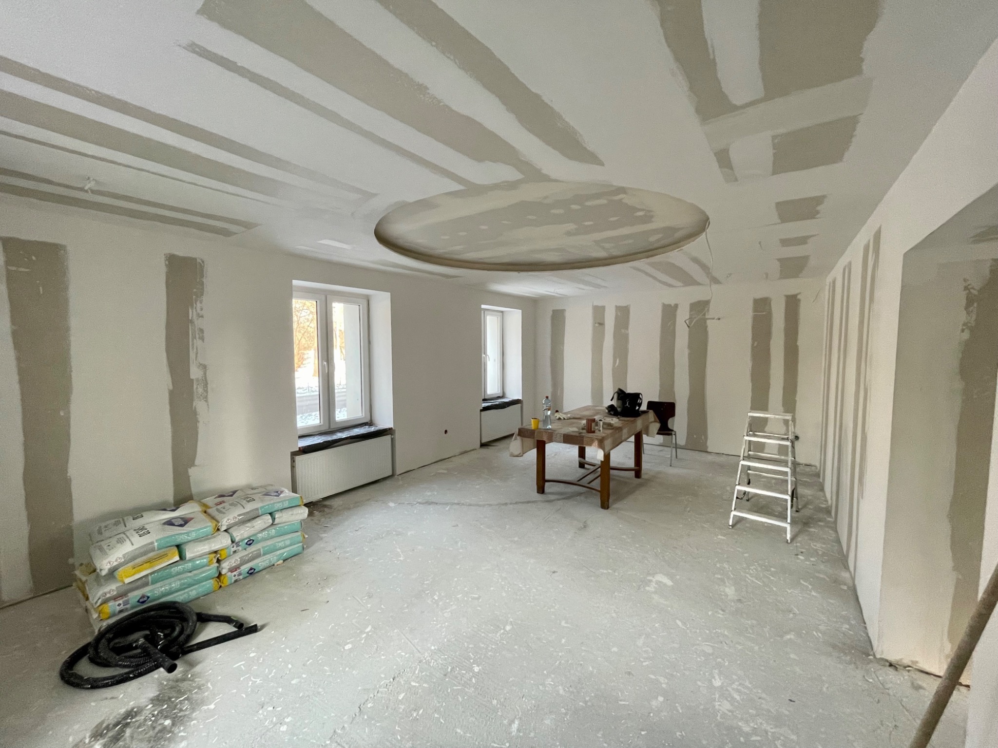 Wnętrze w trakcie remontu: surowy stan ścian z nałożonymi pasami gładzi, worki z zaprawą na podłodze, okrągła dekoracja sufitowa, drabina, stół z narzędziami, dwa okna.