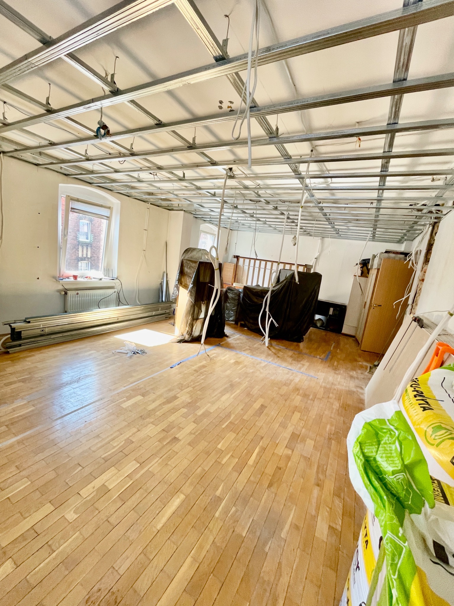 Wnętrze w trakcie remontu: widoczna metalowa konstrukcja sufitu podwieszanego, profile na podłodze, zabezpieczone meble folią, parkiet, okno z widokiem na budynek z czerwonej cegły.