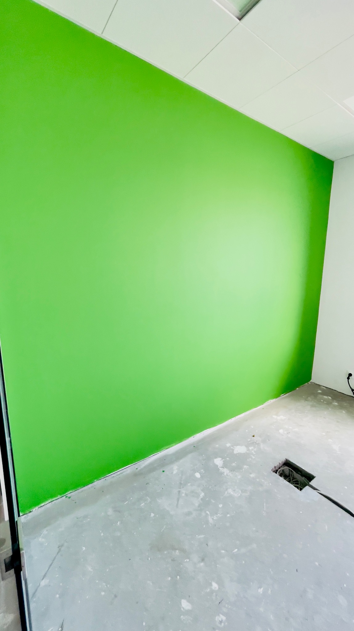 Świeżo pomalowana na zielono ściana w pustym pomieszczeniu z betonową podłogą i otwartym gniazdem elektrycznym w podłodze.