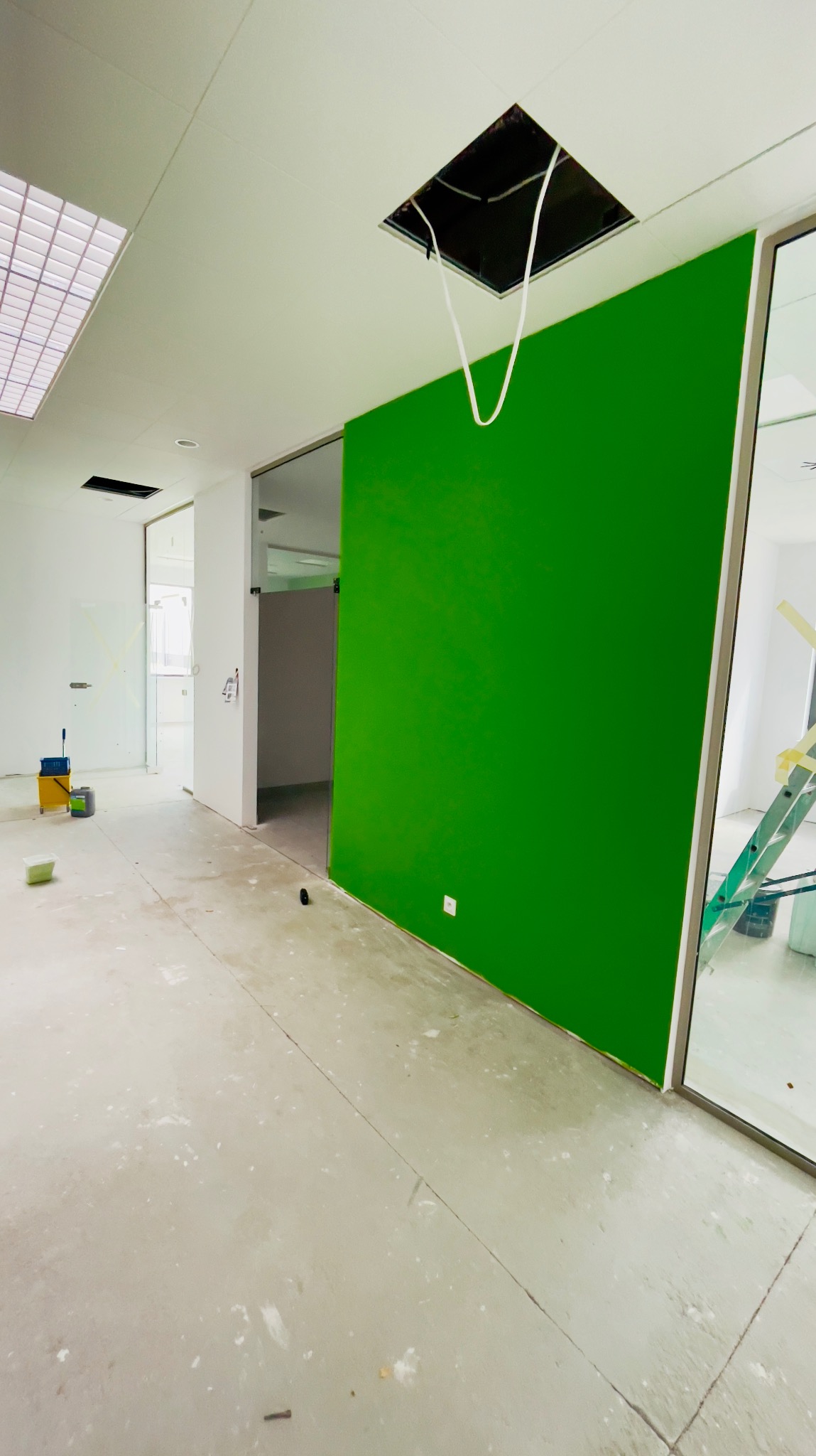 Świeżo pomalowana na jaskrawy zielony kolor ściana w biurze, z widocznymi śladami farby na podłodze, otworami w suficie i drabina oparta o szklaną ścianę.