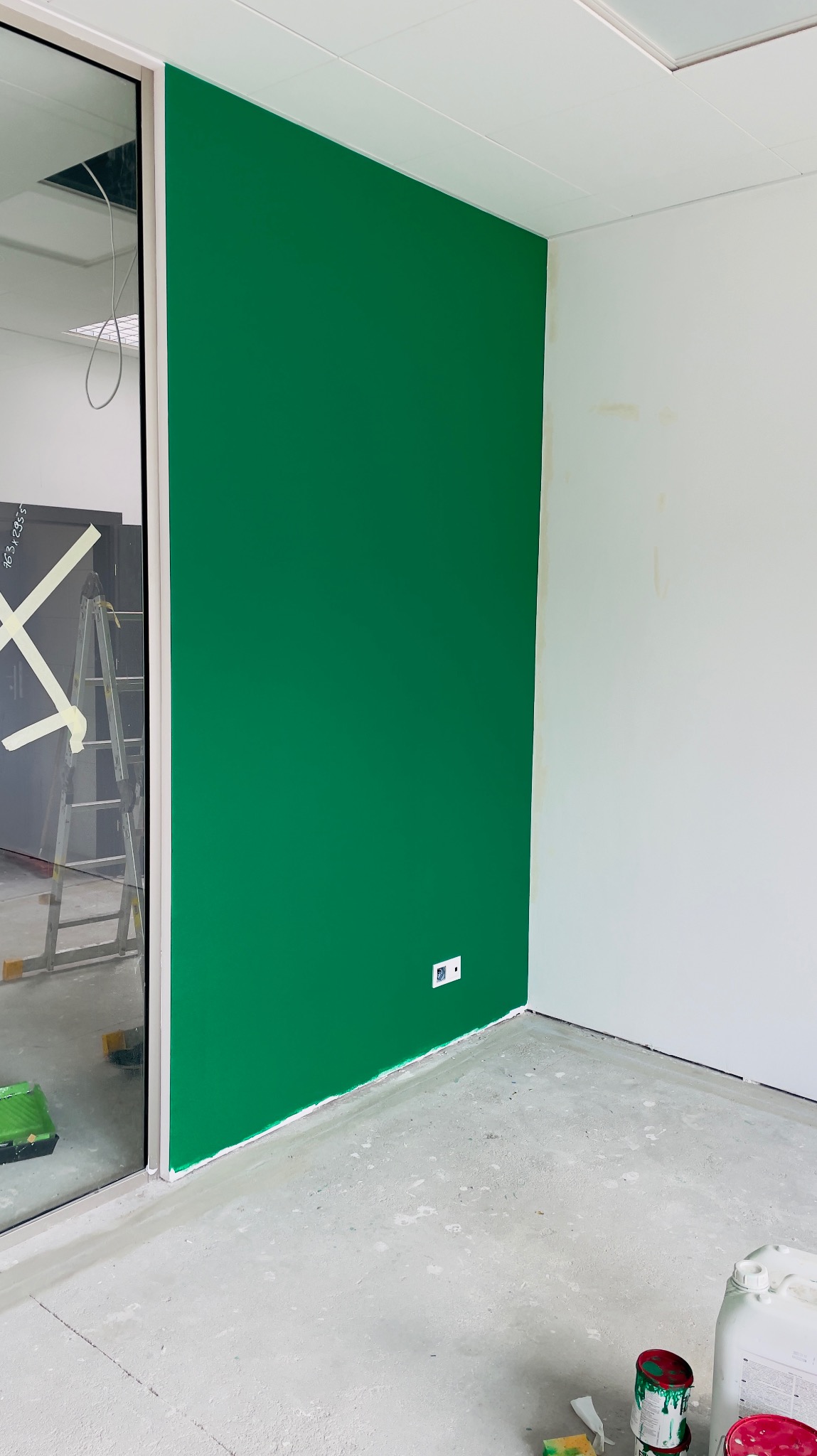 Świeżo pomalowana na intensywny zielony kolor ściana w pomieszczeniu biurowym, widoczne gniazdko elektryczne, fragment drabiny malarskiej i akcesoria malarskie na podłodze, surowy beton na podłodze.