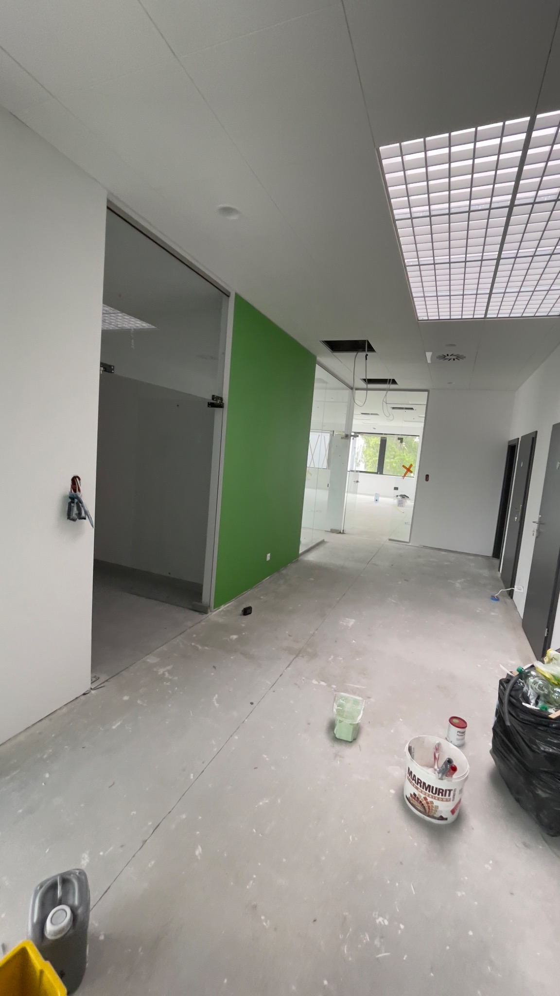 Wnętrze biurowe w trakcie malowania: świeżo pomalowana zielona ściana, widoczne puszki z farbą i ślady farby na podłodze, szklane ścianki działowe.