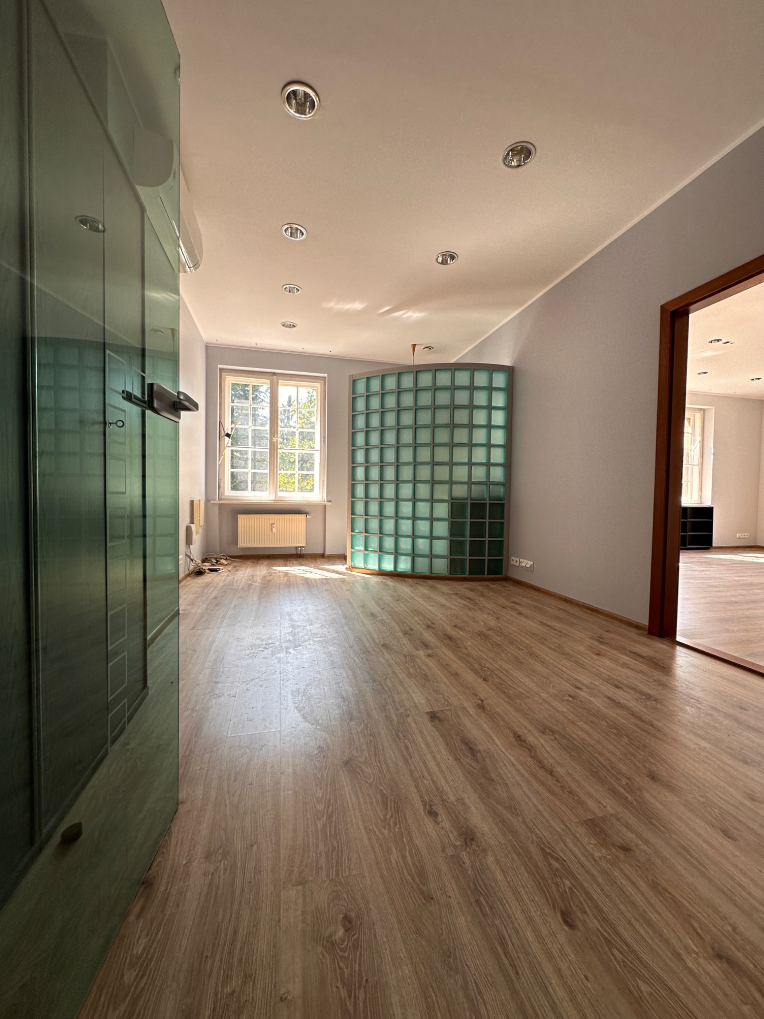 Widok wnętrza z nową podłogą laminowaną, szklaną ścianką działową z luksferów, świeżo pomalowanymi ścianami i widocznym grzejnikiem pod oknem.