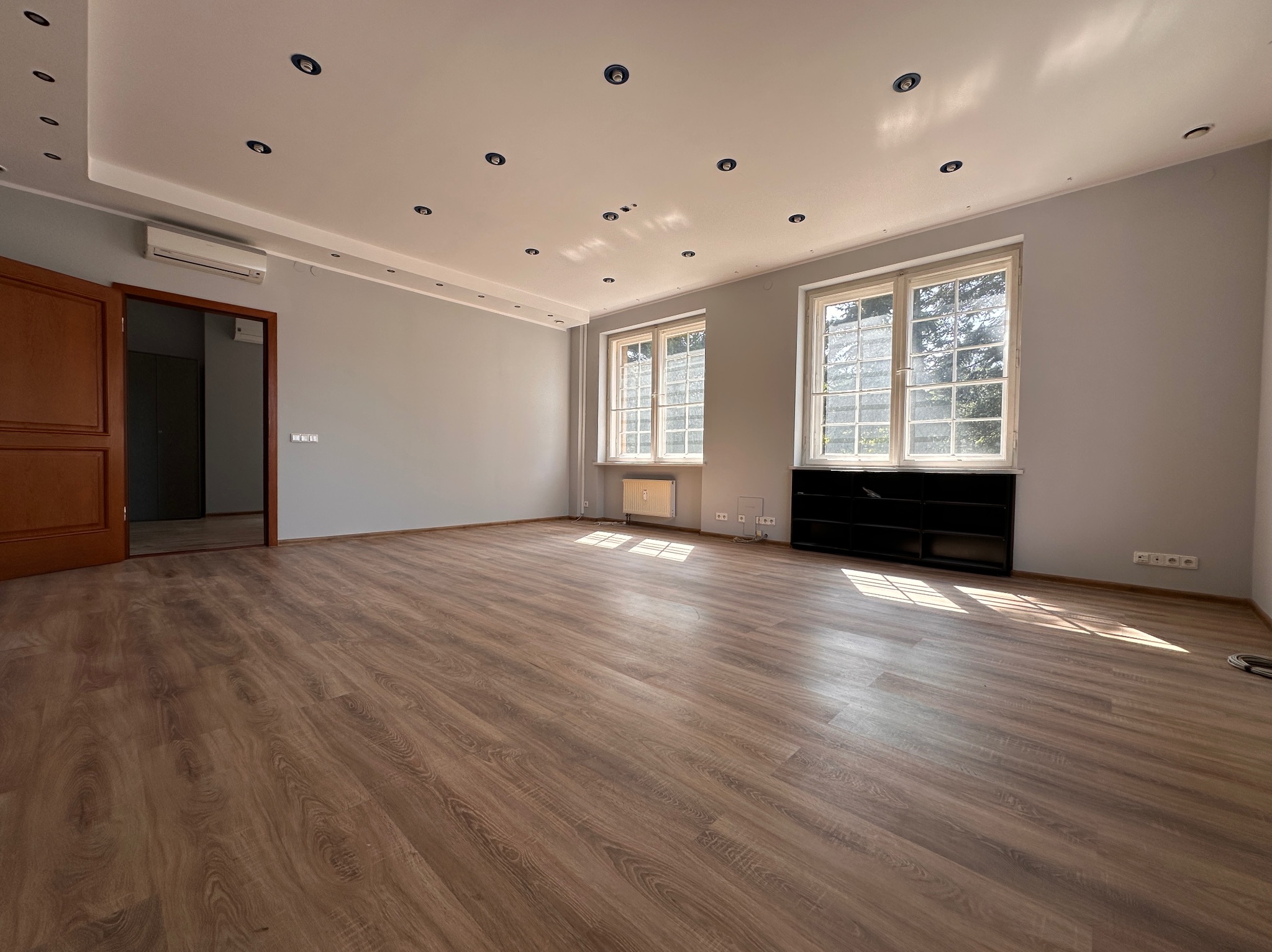 Puste, jasne pomieszczenie z drewnianą podłogą, szarymi ścianami, dwoma oknami z kratami i otwartymi drzwiami. Widoczne punkty świetlne w suficie i klimatyzator.