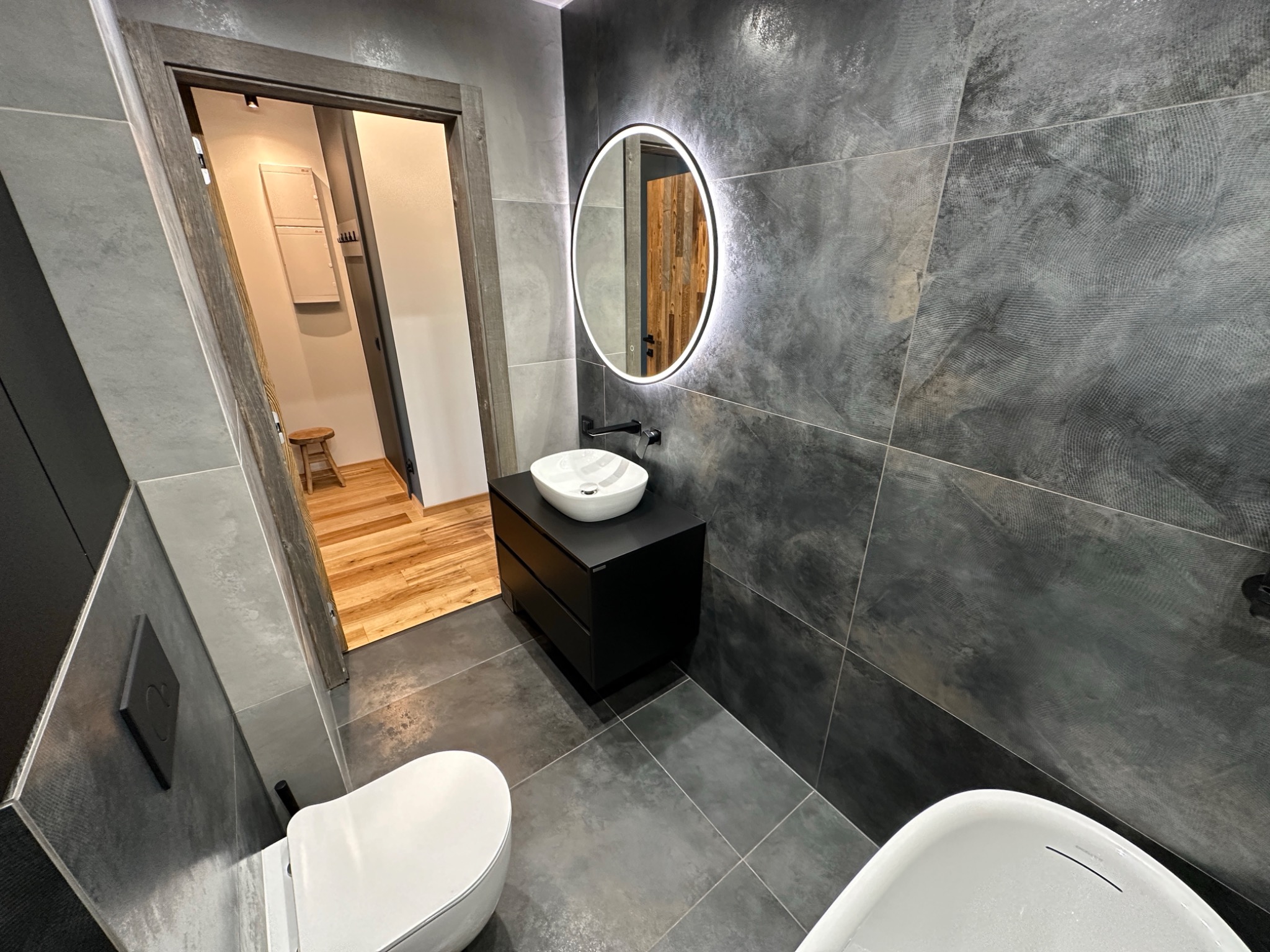 Nowoczesna łazienka z szarymi płytkami imitującymi beton, okrągłym lustrem z podświetleniem LED, białą umywalką nablatową na czarnej szafce i częściowo widoczną wanną oraz toaletą.