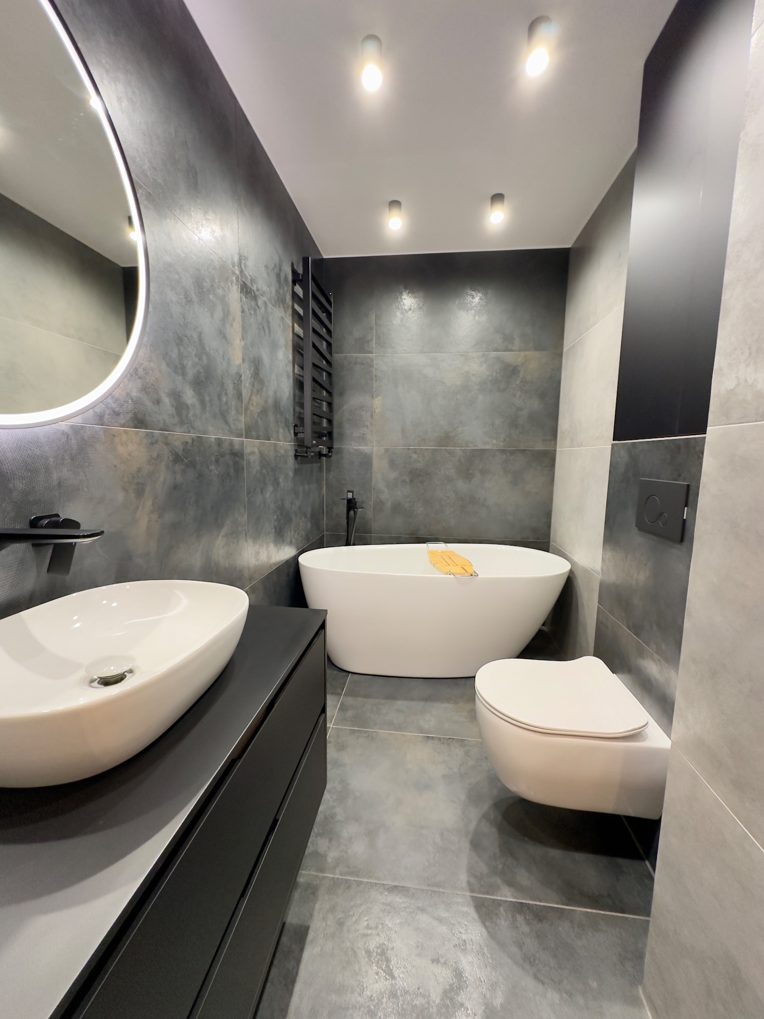 Nowoczesna łazienka w Katowicach z wolnostojącą wanną, umywalką nablatową na czarnej szafce i wiszącą toaletą, utrzymana w szaro-czarnej tonacji z okrągłym lustrem z podświetleniem LED.