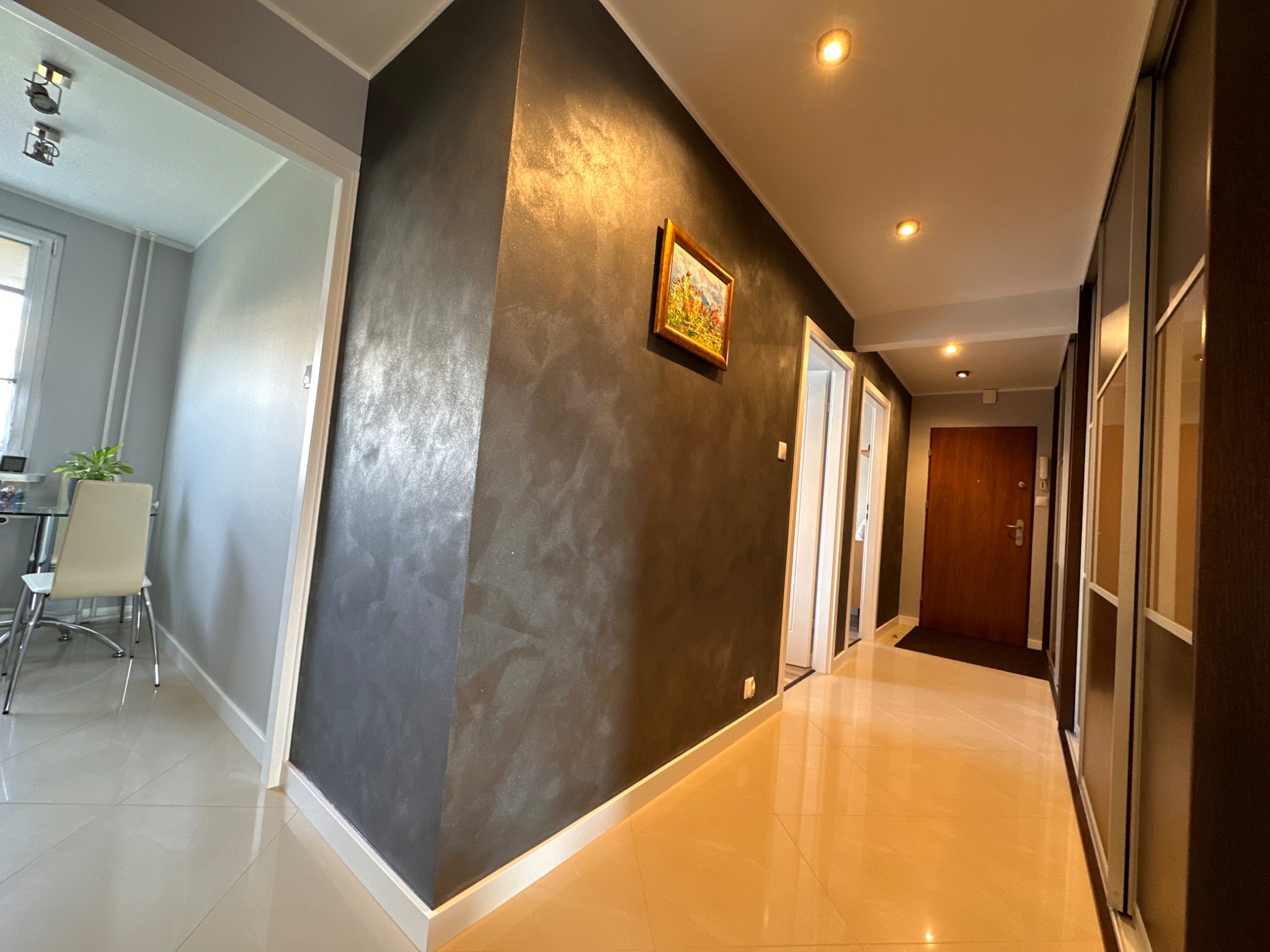 Korytarz z dekoracyjną, ciemną ścianą z efektem metalicznego połysku, obraz w złotej ramie, jasne płytki podłogowe i widok na sąsiednie pomieszczenia przez otwarte drzwi.