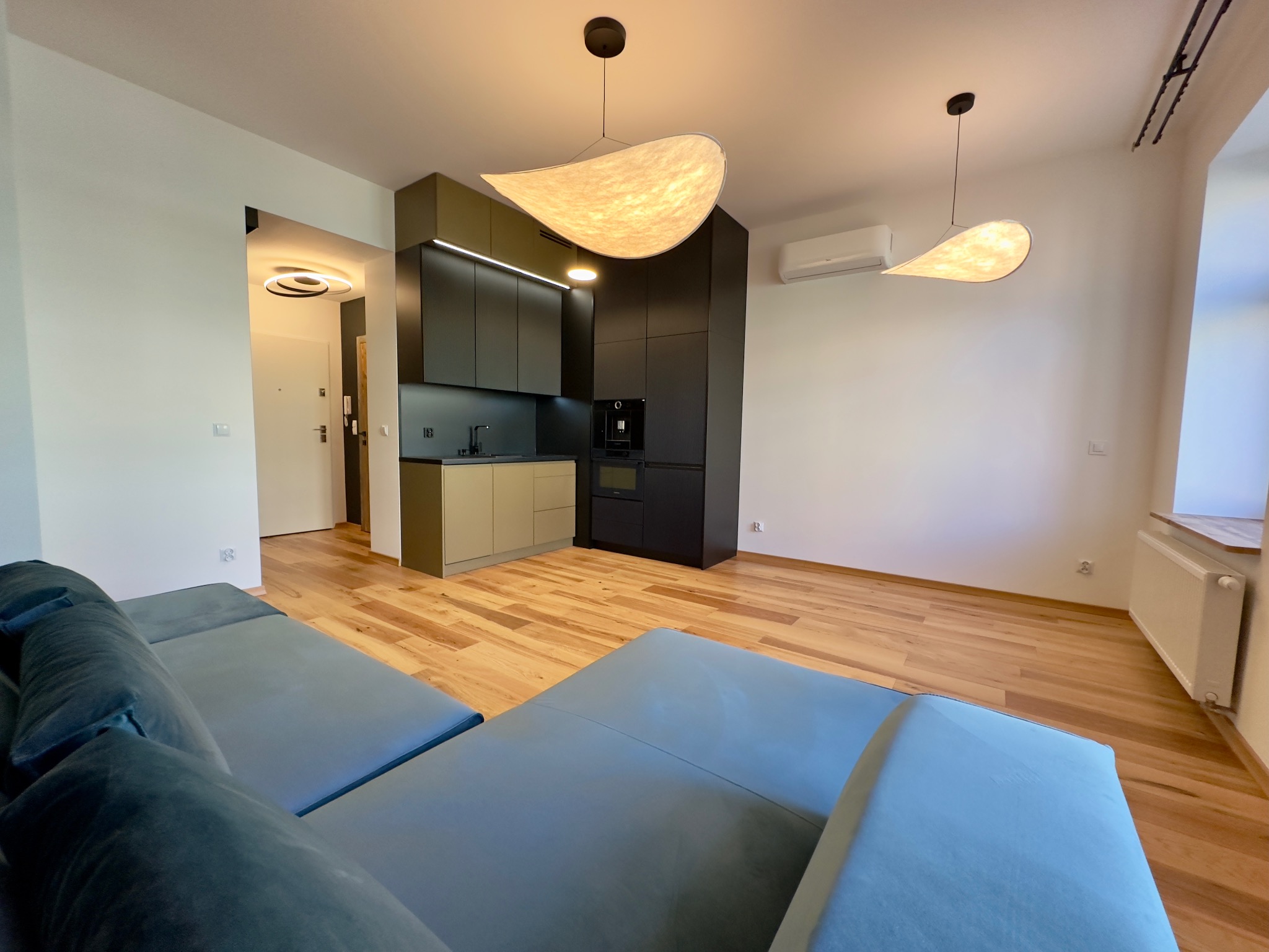 Nowoczesna kuchnia z zabudową w kolorach oliwkowym i czarnym, oświetlona wiszącymi lampami o organicznym kształcie, widok z perspektywy salonu z szarą sofą.