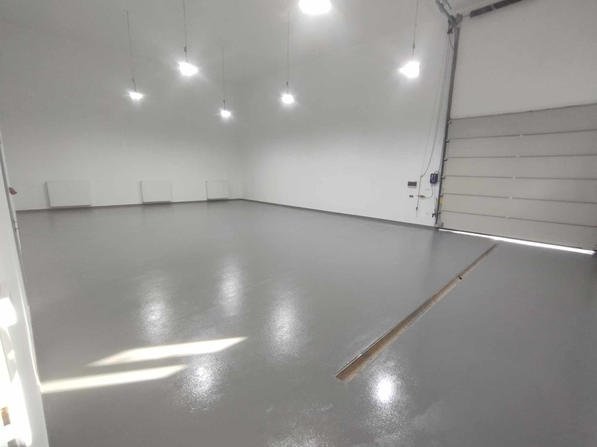 Szara, błyszcząca posadzka w hali przemysłowej z widocznym odpływem liniowym i oświetleniem sufitowym.