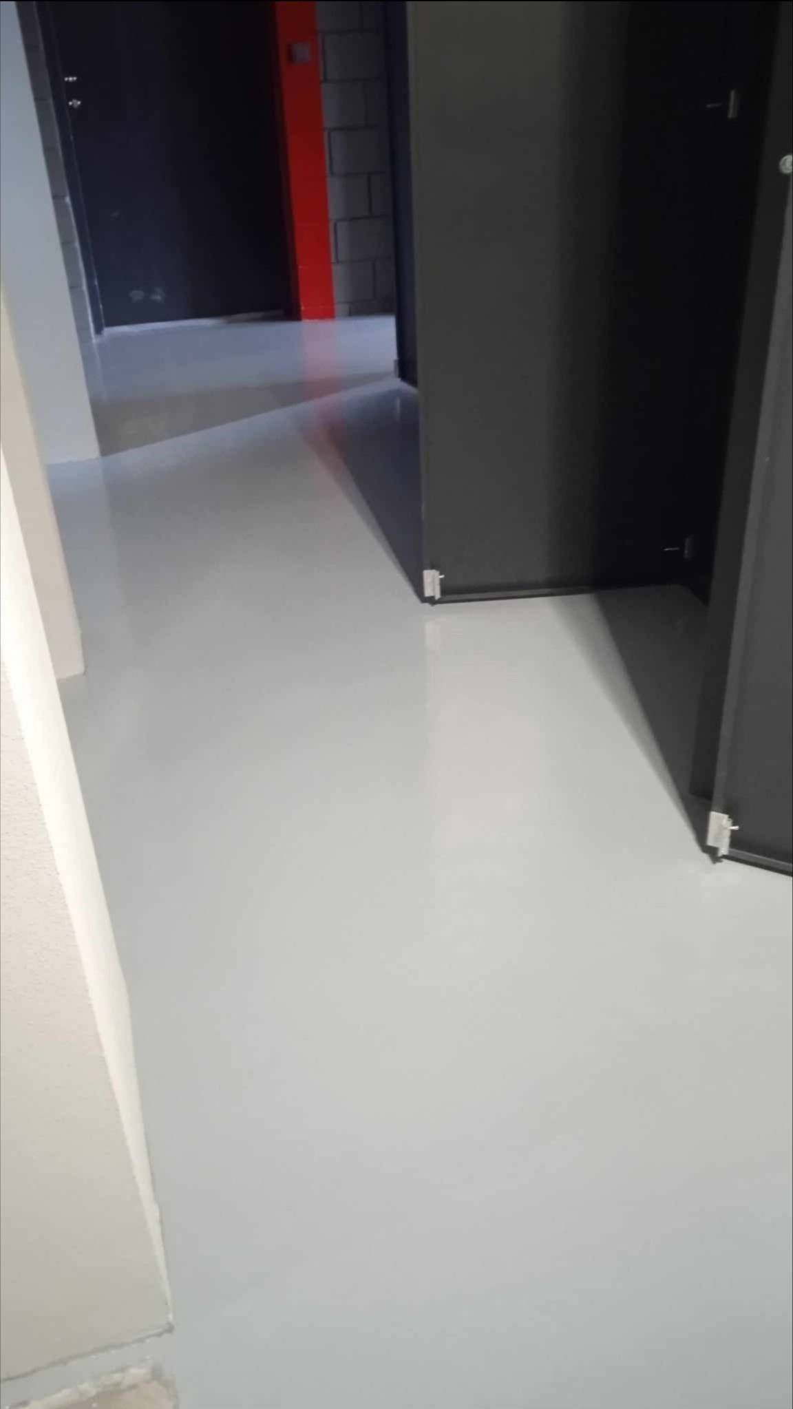 Jasnoszara, gładka posadzka żywiczna w korytarzu z ciemnymi drzwiami i fragmentem ściany z cegieł w tle.