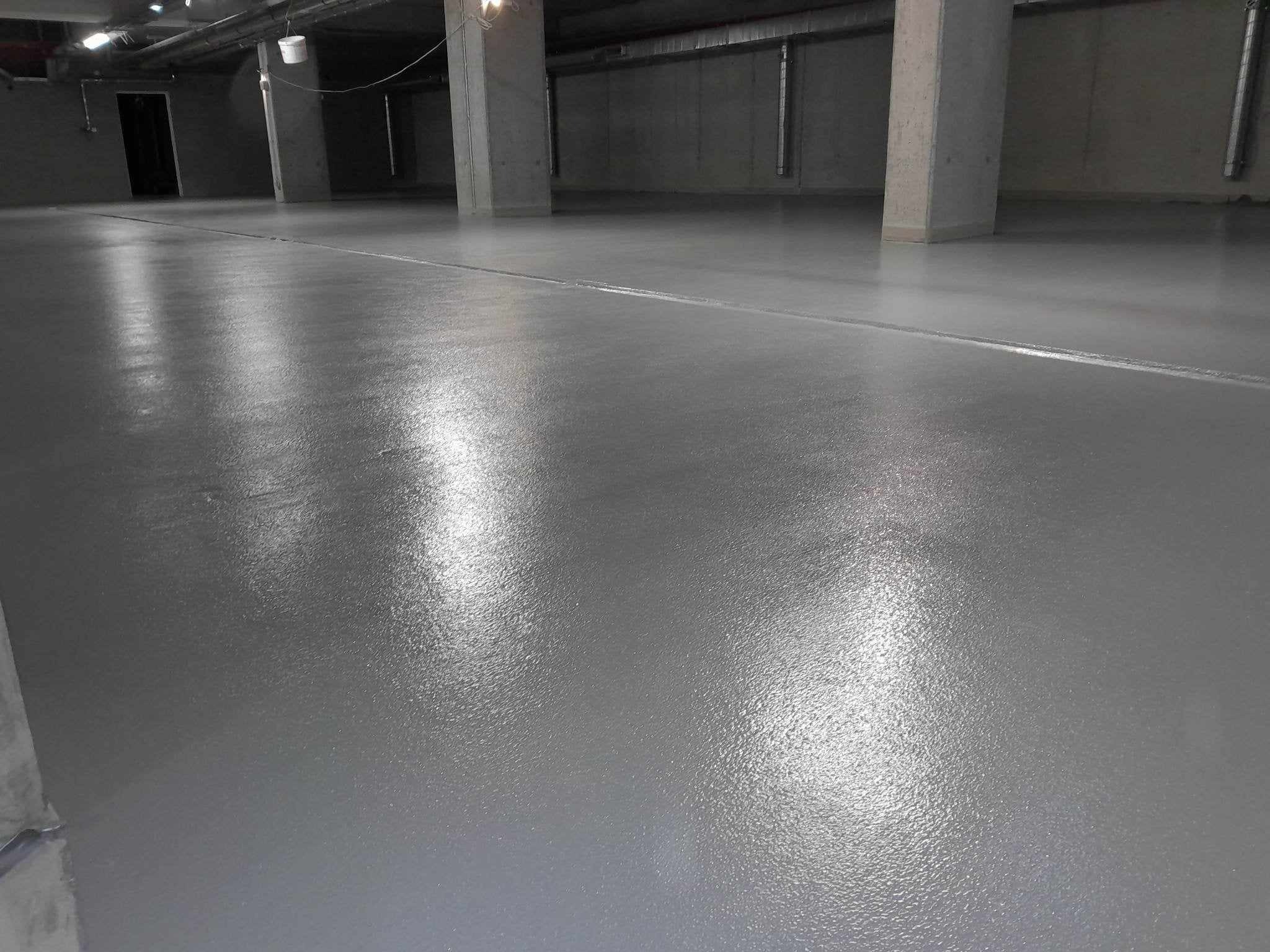 Gładka, szara powierzchnia posadzki w hali przemysłowej, z widocznymi betonowymi kolumnami i rurami pod sufitem, oświetlona sztucznym światłem.