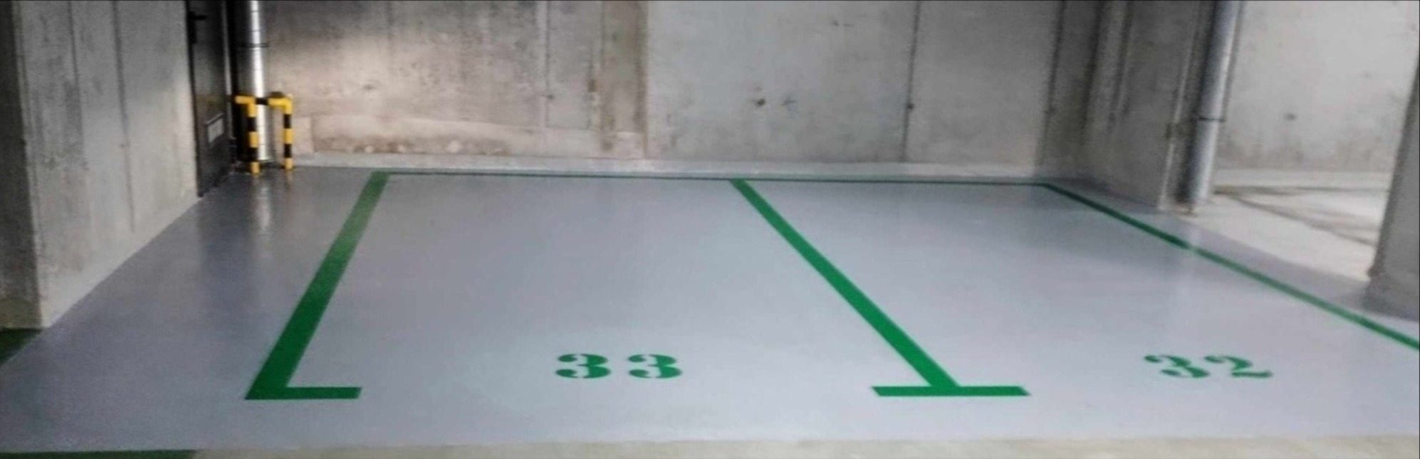 Oznakowane, jasnoszare miejsce parkingowe w garażu podziemnym z numerami 32 i 33, wykończone zielonymi liniami na gładkiej powierzchni.