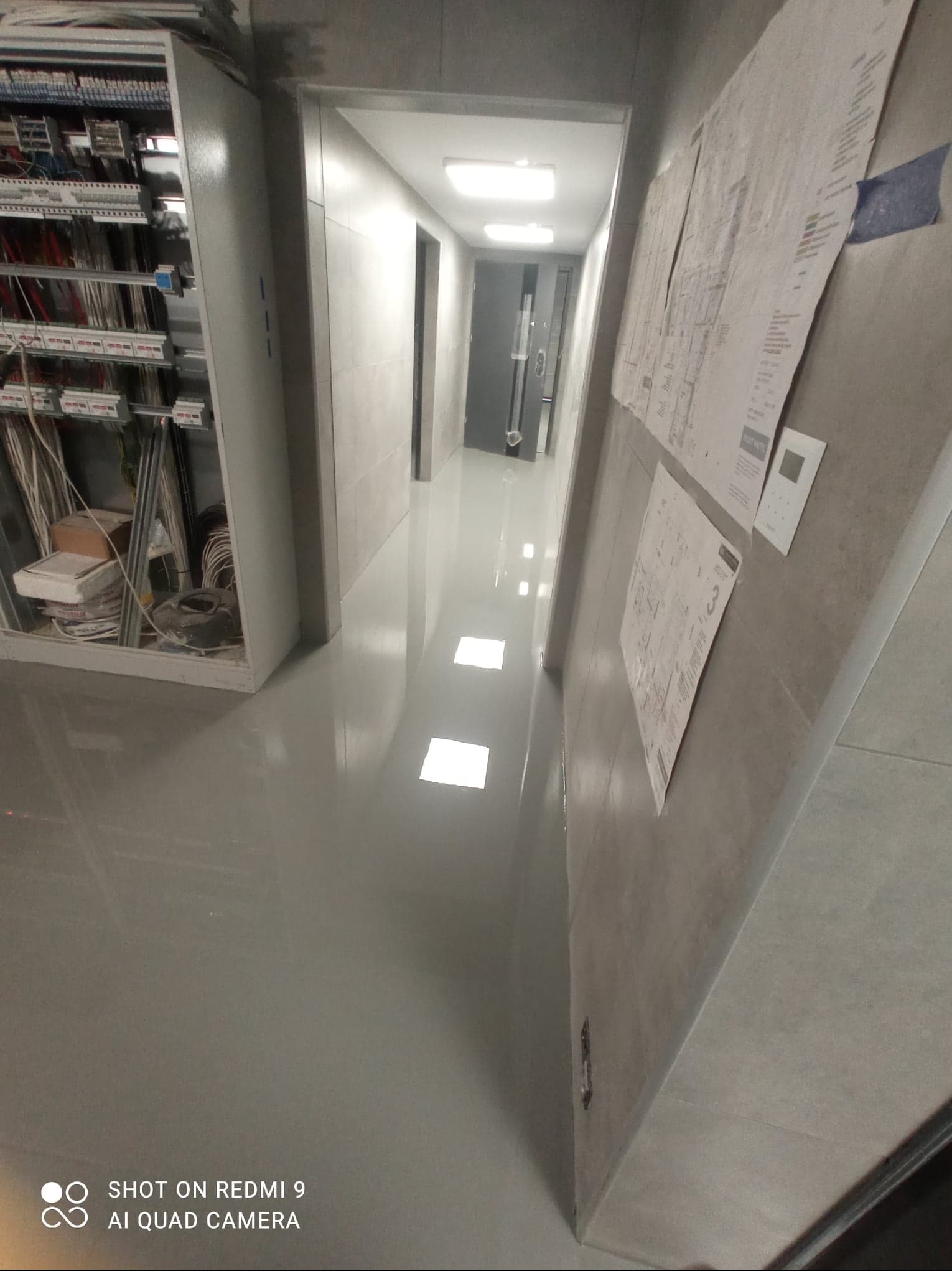 Korytarz w budynku przemysłowym z szarą, błyszczącą posadzką. Na ścianie widoczne plany techniczne przyklejone taśmą. Po lewej stronie szafa elektryczna z okablowaniem.