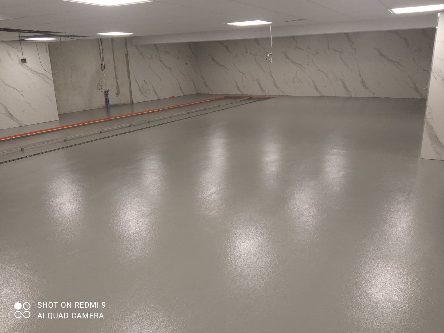 Szara, gładka posadzka w przestronnym pomieszczeniu z marmurowymi panelami ściennymi. Widoczne pomarańczowe rury i metalowe elementy w podłodze.