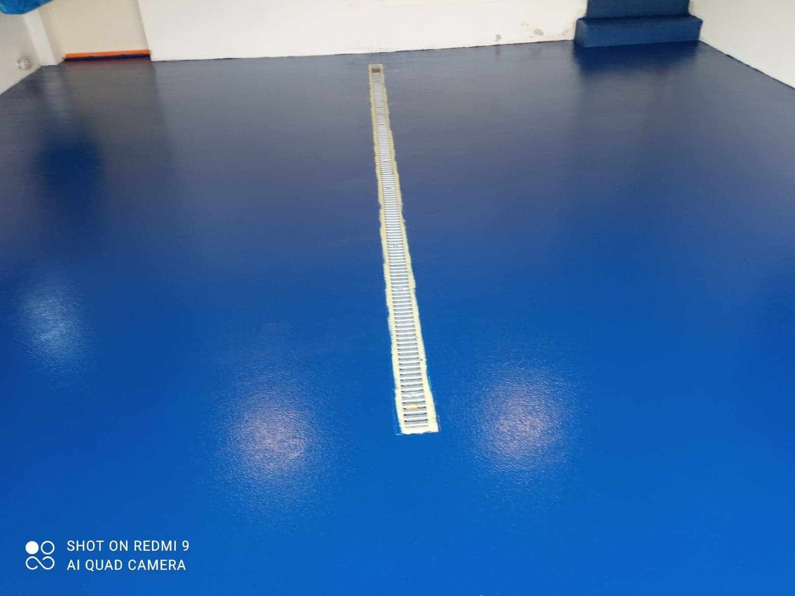 Gładka, niebieska powierzchnia podłogi z żywicy z liniowym odpływem podłogowym pośrodku, widoczne odbicie światła.