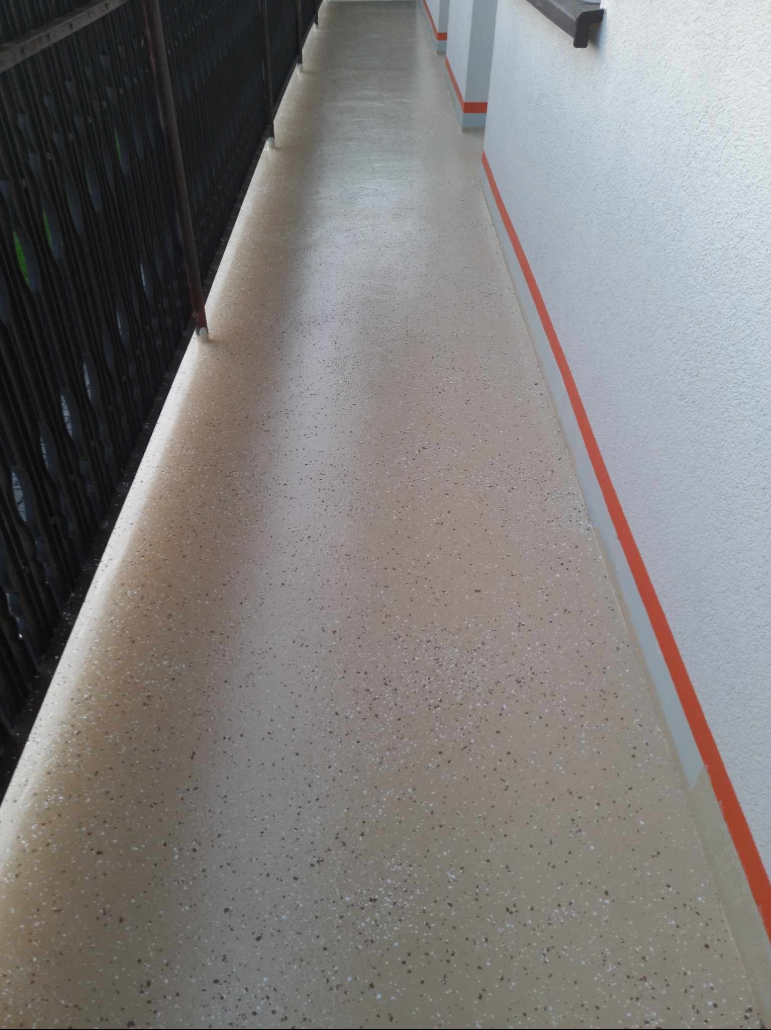Długi balkon z nową, beżową posadzką z drobnymi, ciemnymi i jasnymi plamkami, wykończoną pomarańczowym paskiem przy ścianie. Balustrada z ciemnego metalu po lewej stronie.
