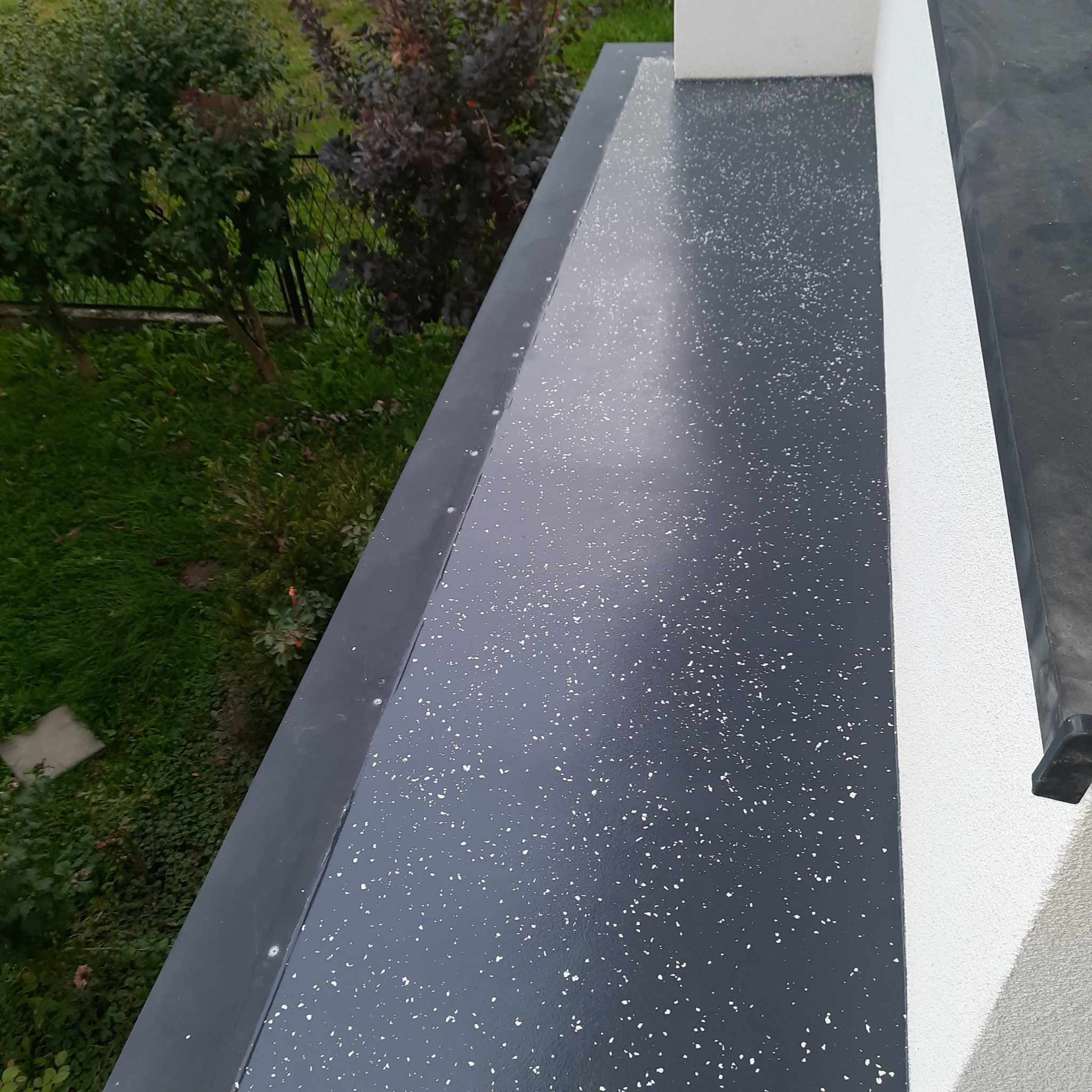 Szara powierzchnia balkonu wykończona żywicą z białymi drobnymi wtrąceniami, widok z góry na zewnątrz, z fragmentem zieleni w tle.