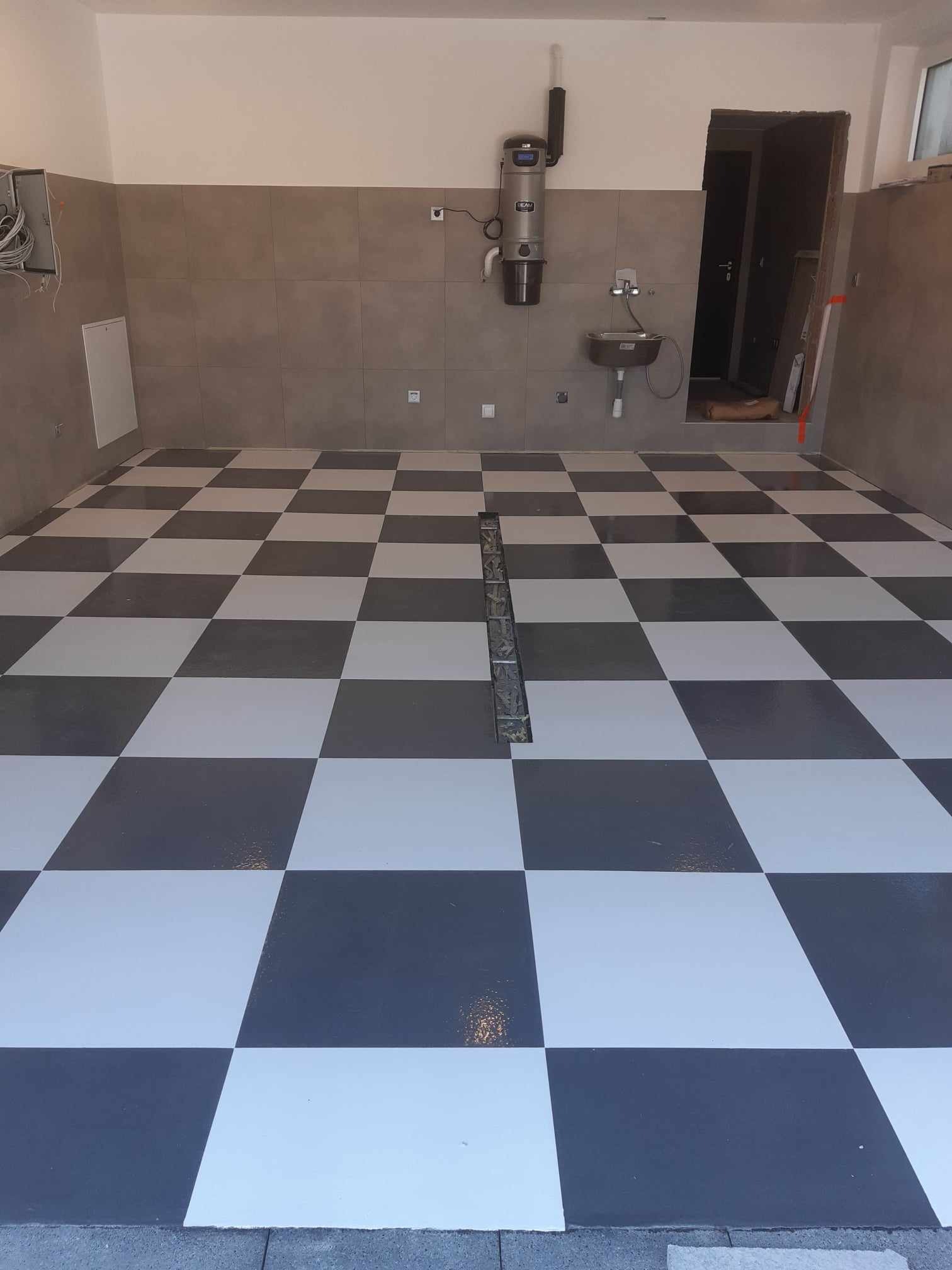 Wnętrze garażu lub pomieszczenia gospodarczego z posadzką w szachownicę, wykonaną z ciemnoszarej i białej żywicy. Widoczny odpływ liniowy na środku posadzki, umywalka ze stali nierdzewnej...