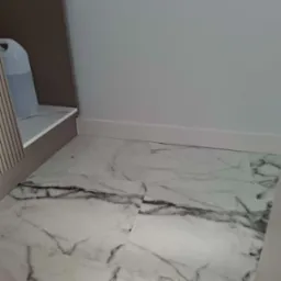 Ułożone białe panele podłogowe imitujące marmur z widocznymi szarymi żyłami, biała listwa przypodłogowa, fragment szafki z beżowymi lamelami i przezroczystym kanistrem.