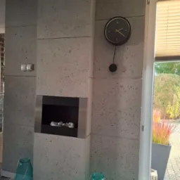 Ściana wyłożona betonowymi płytami z wbudowanym biokominkiem i wiszącym zegarem, dekoracyjne lampiony na podłodze, widok na ogr&oacute;d przez okno.