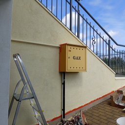 MajsterReda - Ściana budynku po malowaniu z żółtą skrzynką gazową, widoczne narzędzia malarskie i drabina. Świeżo pomalowana elewacja w ciepłym odcieniu beżu.